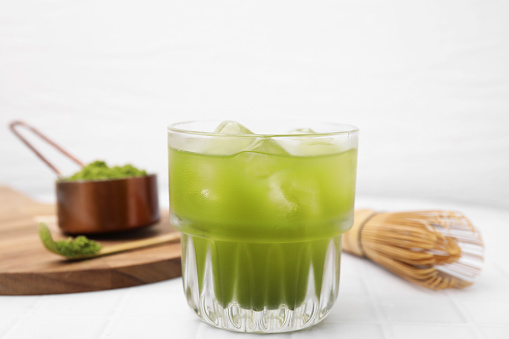Glass of delicious iced green matcha tea, powder and bamboo whisk on white tiled table, closeup