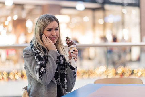 Beautiful blonde woman sitting in shopping mall and having toothache after eating ice cream.