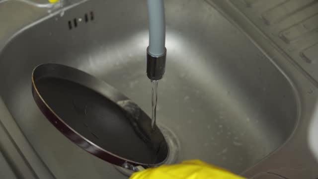 A woman in yellow gloves washes a frying pan, rinses off the detergent with water.