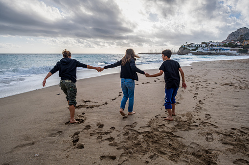 Rear view of a young mother and her teenage sons enjoying time together along a deserted beach, holding hands and walking on a cloudy day.