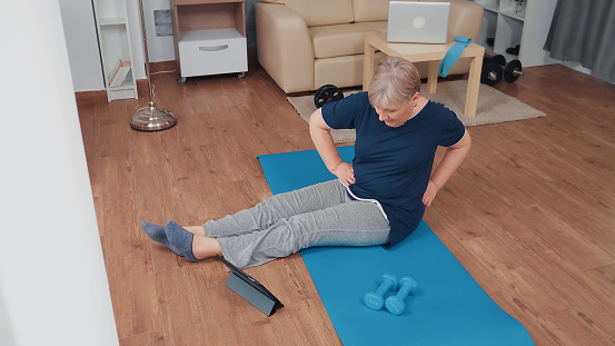 Senior woman doing warm up exercise during a video call with personal trainer. Old person pensioner online internet exercise training at home sport activity at elderly retirement age