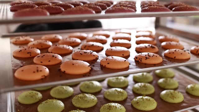 Chef placing a tray full of colorful macarons on a metal shelf with another macaroons, situated in a bakery. Process of making delicious french dessert. Confectionery in the pastry workshop.