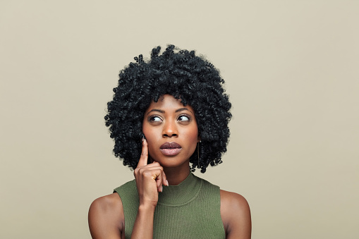 Headshot of pensive black young woman with afro hairstyle wearing green top standing against beige background and looking away with hand on chin.