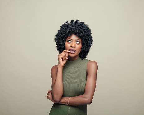 Pensive black young woman with afro hairstyle wearing green top standing against beige background and looking up with hand on chin.