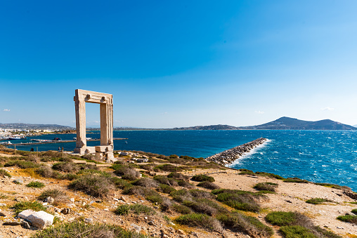 In the warm familiarity of a Naxos afternoon, an iconic snapshot captures an extraordinary tableau. The Apollo Gate stands grandly, a silent testament to the past, its glory gently caressed by the sun's rays. Nearby, a strategic windbreak deflects the Aegean Sea's playful swells, shielding the serene Naxos harbour. It is a simultaneous portrayal of the island's historic grandeur and its unyielding resilience in the face of nature's caprices.