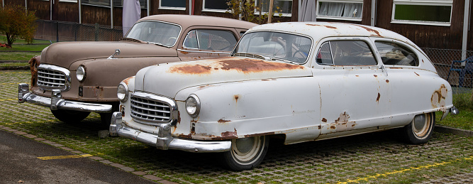 two american cars from the 1950s, old and rusty without paint, brown and white, windows are partly broken with holes, front right photographed, on the day without people