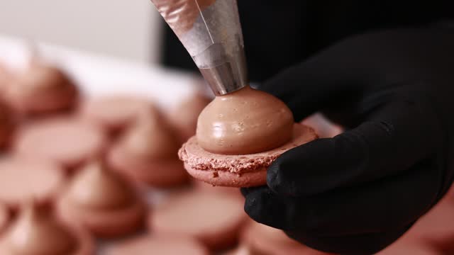 Making french desert macarons. Chef hands in black gloves with confectionery bag add cream to the ready halves of brown chocolate macaroons at pastry shop. cooking, food and baking concept.