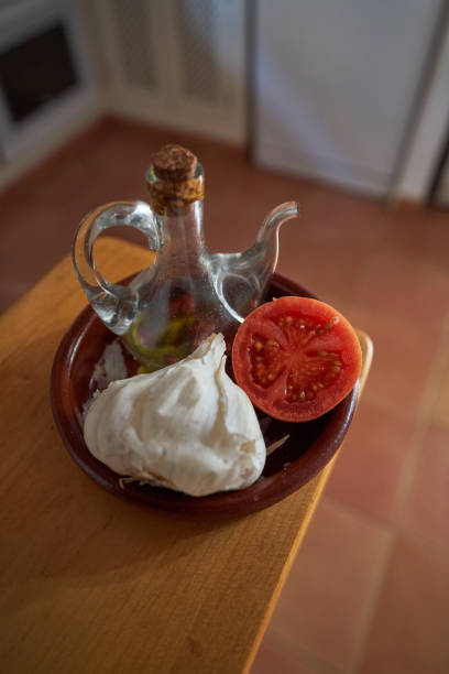 From above head of garlic with half of tomato on plate with bottle of oil in light kitchen during cooking process
