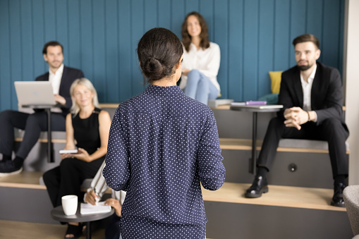 Back view of young female boss talking to diverse employees on corporate meeting, motivating staff, presenting project plan. Business teacher giving educating lecture, seminar