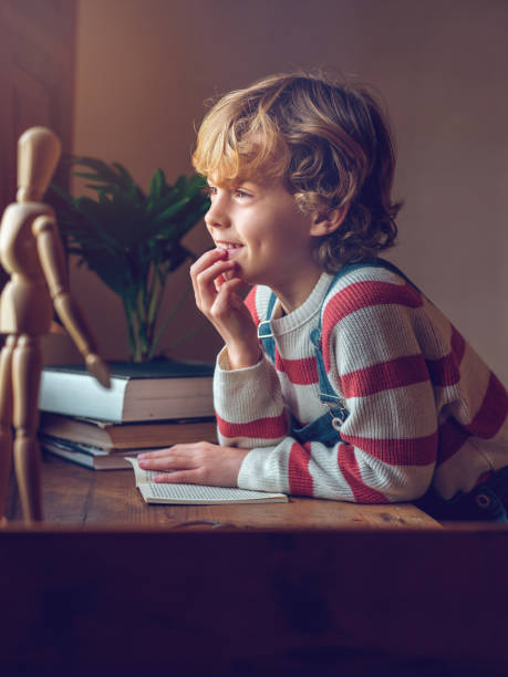 Cute schoolboy with book sitting at table Side view of adorable little boy in sweater sitting at wooden table with books and looking away in dreams Distracted stock pictures, royalty-free photos & images