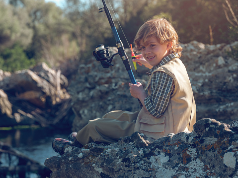 Full body side view of cheerful boy sitting on rocky coast with fishing rod while catching fish near river in nature