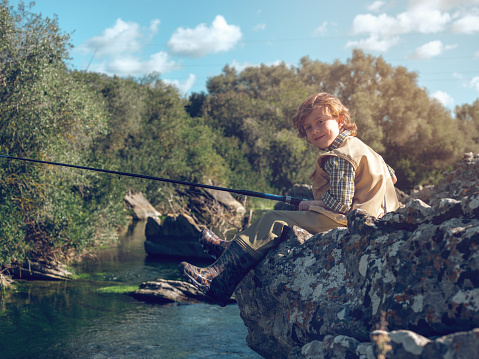 Little smiling child looking at camera while sitting on rocky coast of lake and catching fish with rod in sunlight