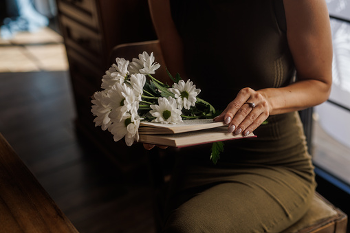 Woman sitting by the window in her home and holding an open book with a bouquet of daisy flowers inside.