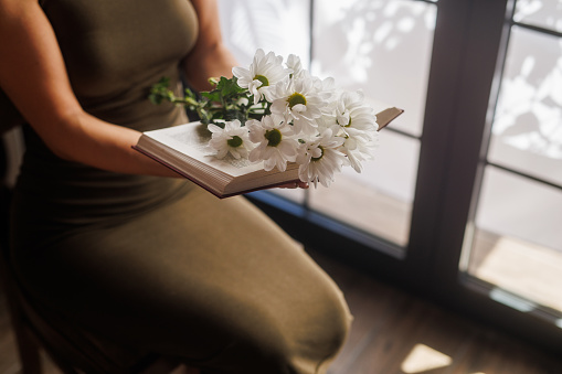 Woman sitting by the window in her home and holding an open book with a bouquet of daisy flowers inside.