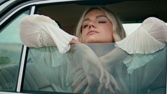 Girl looking through car window close up. Beautiful woman traveler leaning on automobile glass wearing white dress. Relaxed young blonde model posing in retro auto looking outside. Travel trip concept