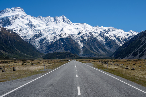 Discover the picturesque country road in New Zealand's South Island leading to the renowned Mount Cook National Park. Behold the stunning sight of snow-capped Mount Cook, majestically framed by other towering mountain peaks.