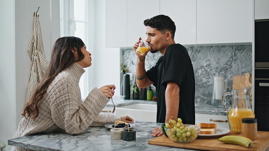 Romantic newlyweds enjoying breakfast at kitchen. Handsome man drinking orange juice glass closeup. Gorgeous woman sitting table chewing food talking. Positive lovers speaking at morning together
