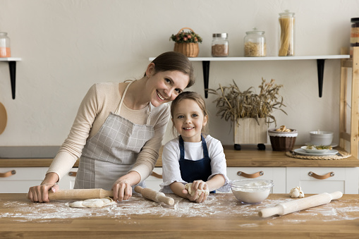 Happy sweet little girl and loving mom in aprons baking in home kitchen, rolling dough on floury messy table, shooting culinary hobby, looking at camera with toothy smiles, posing for family portrait