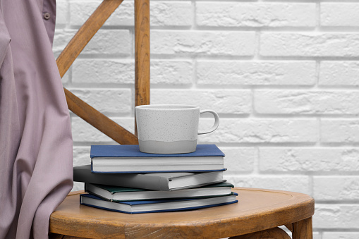 Ceramic cup and stack of books on wooden chair against white brick wall, space for text