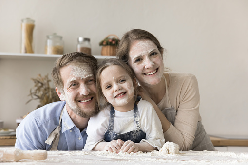 Happy young mom, dad and toddler kid in aprons with funny floury facial masks cooking dessert in home kitchen, having fun, posing at table with heap of flour, looking at camera, laughing