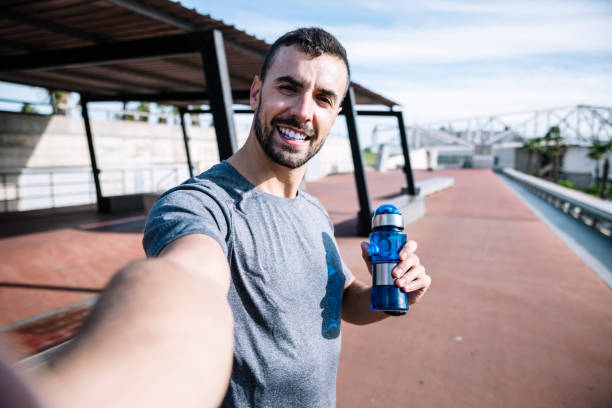 istock Runner man drinking water and taking a selfie after exercise outside. Handsome sports guy holding a bottle taking a picture and looking at camera. 1925421536