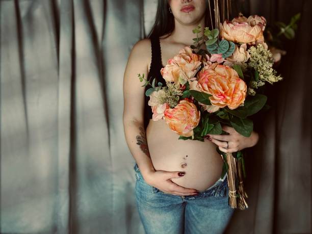 moody portrait of a pregnant young adult woman holding a bouquet of flowers. - pierced abdomen flower beauty - fotografias e filmes do acervo