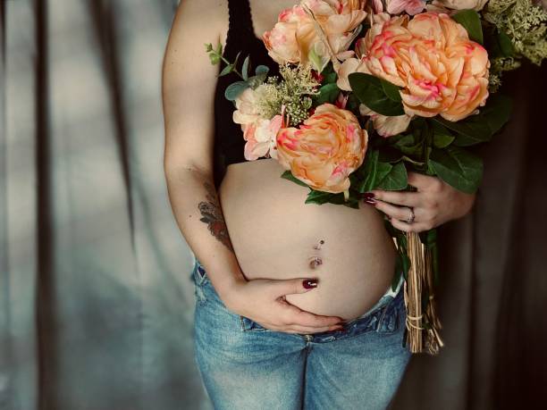 moody portrait of a pregnant young adult woman holding a bouquet of flowers. - pierced abdomen flower beauty ストックフォトと画像