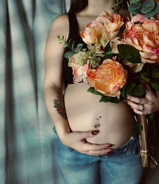 moody portrait of a pregnant young adult woman holding a bouquet of flowers. - pierced abdomen flower beauty ストックフォトと画像