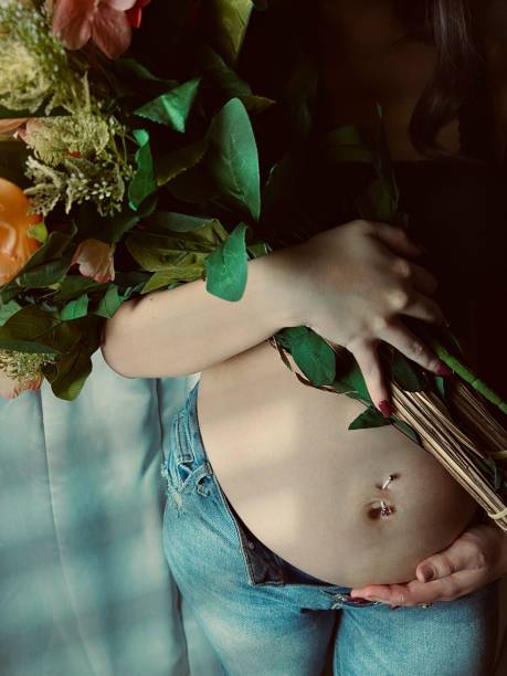 moody portrait of a pregnant young adult woman holding a bouquet of flowers. - pierced abdomen flower beauty ストックフォトと画像