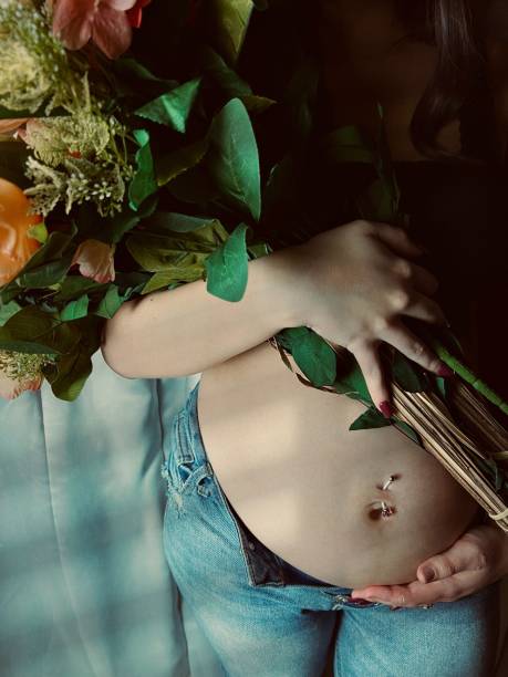 moody portrait of a pregnant young adult woman holding a bouquet of flowers. - pierced abdomen flower beauty - fotografias e filmes do acervo