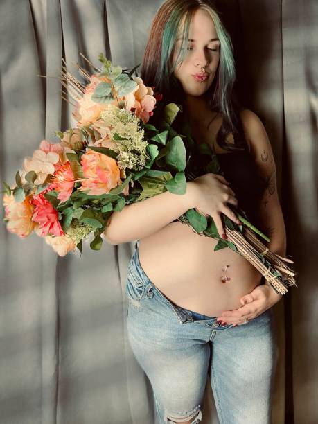 moody portrait of a pregnant young adult woman holding a bouquet of flowers. - pierced abdomen flower beauty - fotografias e filmes do acervo