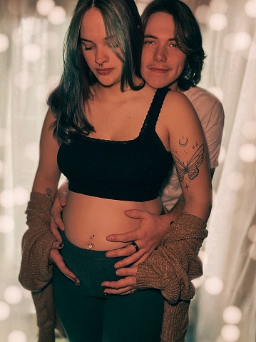 Moody portrait of a young adult/teen couple standing in front of a natural lit white curtained window, holding each other. The women is 7 months pregnant. Bokeh lights and shadows in the background. Part of a series of maternity pictures.