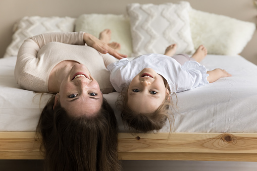 Happy mom and cheerful cute toddler girl upside down heads hanging from mattress. Positive mother and child lying on backs on wooden comfortable double bed together, looking at camera for portrait