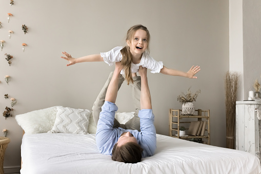 Happy dad lying on back, holding joyful carefree little daughter girl in arms, lifting child up in air for playing airplane, laughing, having fun, enjoying leisure, fatherhood, practicing acroyoga