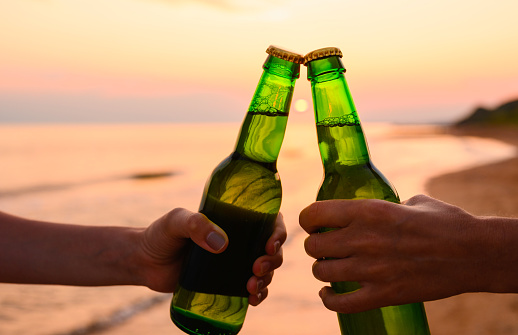 Concept of meeting friends on vacation. close-up of a bottle of beer in hands against the backdrop of sunset and sea. Selective focus, free space