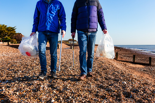 Two volunteers collect plastic waste that has been washed up on the beach. The two people are carrying transparent bags in which they collect the waste, and grabbers to remove the plastic from the beach.