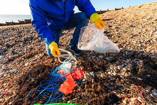 Color image depicting a mid adult man cleaning a beach by picking up plastic waste that has been washed up on the beach.