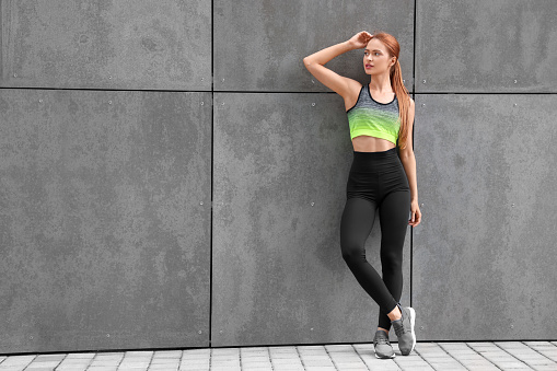 Beautiful woman in stylish gym clothes posing near grey wall on street, space for text