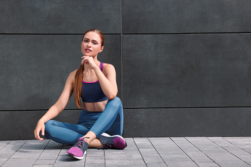 Beautiful woman in stylish gym clothes sitting near dark grey wall on street, space for text