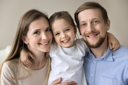 Cheerful sweet little girl hugging young mom and dad with love, tenderness. Happy couple of parents and cute toddler kid looking at camera, smiling, laughing, posing at home for family portrait