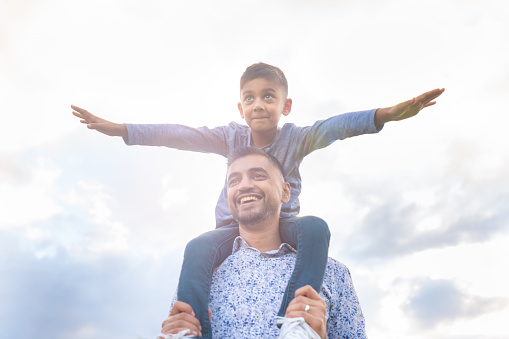 Father carrying his son on his shoulders in the park. The boy has his arms out like an airplane. Both are happy and smiling.