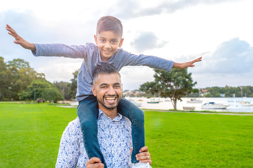 Father carrying his son on his shoulders in the park. The boy has his arms out like an airplane. Both are happy and smiling.