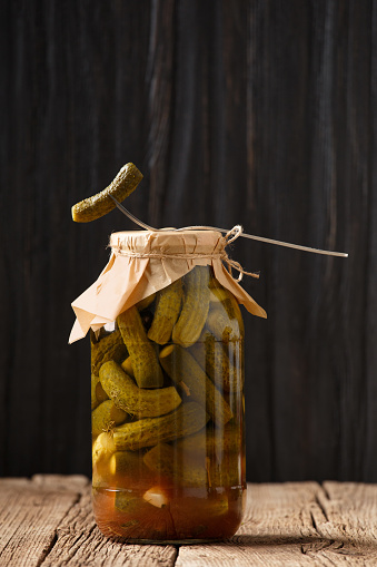 Canned cucumbers in a jar, one pickled gherkin is pricked on a fork and lies on the lid of the bottle, dark wooden background, copy space.
