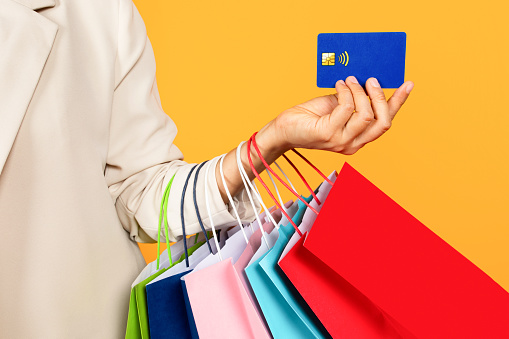 Young asian woman hold many bags on hand, shows credit card, isolated on yellow studio background, cropped. Sale, finance and shopping recommendation, ad and offer, pay and buy purchases