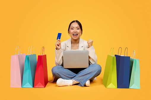 Glad excited millennial chinese lady with shopping bags, credit card, laptop, enjoy sale and win, isolated on orange studio background. Finance for purchases, shopaholic emotions, cashback