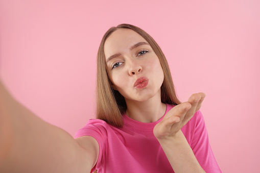 Young woman taking selfie and blowing kiss on pink background