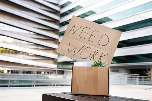 Unemployment Concept. Placard With Need Work Text Put In Box With Personal Belongings Standing On Bench Outdoors Against Modern Business Center Background, Creative Banner For Joblessness Problem