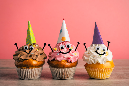 A row of party cupcakes with party hats and googly eyes smiling with their hands in the air.