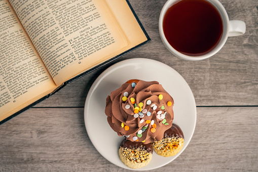 An overhead view of a plate with a cupcake and cookies and a cup of hot tea with an open bible on a table top.