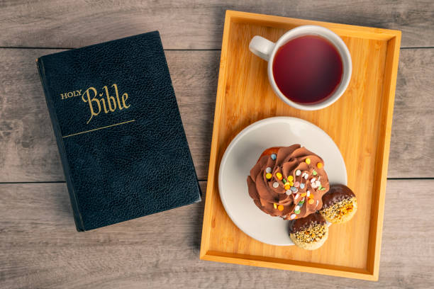 An overhead view of a Holy Bible on a table with a serving tray with hot tea and a cupcake and cookies.